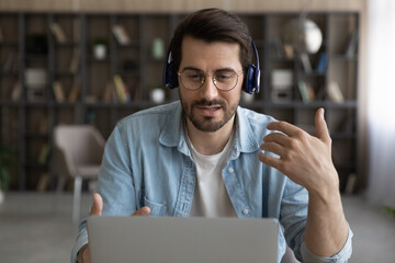 Millennial Caucasian man in earphones look at laptop screen talk on video call on gadget. Young...