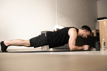 Fototapeta na wymiar Plank it Training at home. Confident young man wearing black sportswear and doing plank position while exercising on the floor in light loft interior. Hard workout.