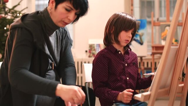 An art teacher assists a child in an art school. The boy makes a drawing sitting in front of a malbert. They are discussing work. Finger holding technique.