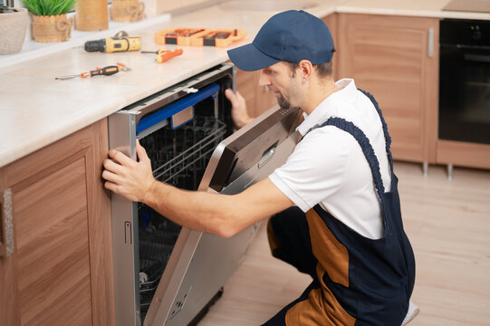 A man or service worker in special clothing installs, disassembles or performs maintenance of a dishwasher built into the kitchen furniture