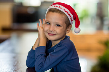 Portrait of a cute little child boy wearing Christmas Santa hat sitting by the table and smiling. Merry Christmas and happy holidays