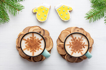 Two mugs of latte decorated with snowflakes near gingerbread mittens on white wooden background