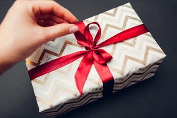 Woman opens Christmas and New year gift box wrapped in paper and decorated with red ribbon bow on black background.