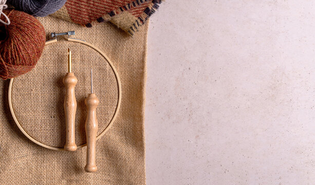 Composition with copy space of punch needle tools on a concrete surface. A jute fabric in an embroidery hoop, two different size of needles and wool balls.
