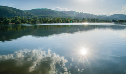 Słońce, chmury, góry i las odbijające się w tafli wody jeziora.