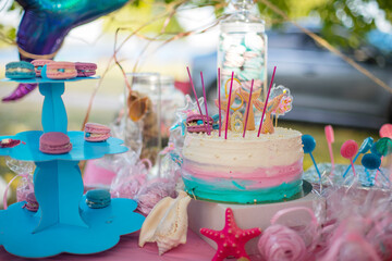 Mermaid Candy Bar. A beautiful cake decorated with mermaid tails.White shell plate full of chocolate candies with blue icing, cookies with shells, stars on the table with shining cloth. 