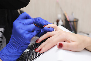 Master in protective gloves during a manicure at beauty salon. Master manicurist varnishes the marsala gel on the nails of a female client. The concept of beauty and health.