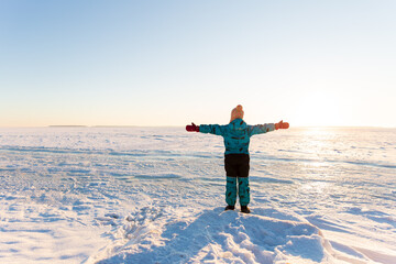 A child in a winter overalls stands with his arms outstretched against the background of a beautiful winter landscape, snow and a clear blue sky. Freedom concept, look ahead. Travel.