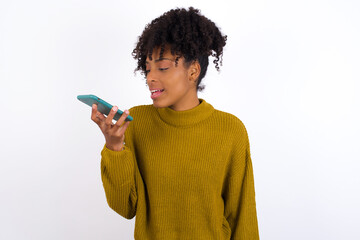 Fototapeta na wymiar Smiling Young beautiful African American woman wearing knitted sweater against white wall, sending voice message on her smart phone. Communication and new technologies concept.