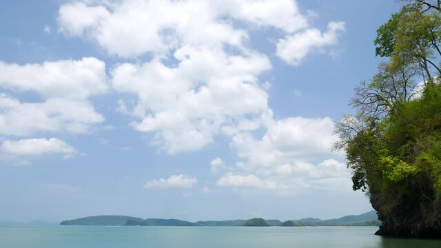 Light white clouds fly and curl, calm tropical sea and small cliff covered with green plants on side. Time lapse shot at hot day, small boats rush across water at distance