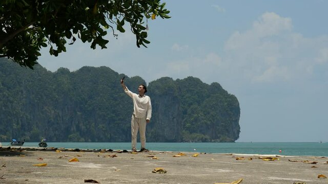 Man take selfie photograph against sea, stand at old pier or quay. Green cliffs of Ao Nang seen on background, hot sunny weather at noon, refreshing breeze from water
