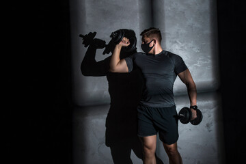 Male athlete wearing protective face mask and training with dumbbell in gym. Coronavirus world pandemic and sport theme.