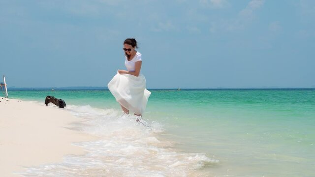 Clear and warm sea water and fine sand, happy woman enjoy beach at tropical island. She stand in splashing waves and turn, laugh and wave long skirt, slow motion shot