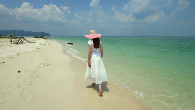 Tourist woman stroll at beautiful beach on tropical island, camera follow behind. Crystal turquoise water of sea slowly washing soft fine sand. Hot sunny weather at Thailand