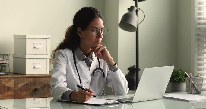 Young female doctor in white coat working on laptop in modern hospital office room, therapist typing on computer consult patient online, makes research, noting, takes useful information from internet