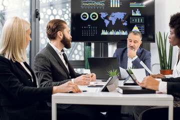 Team of multiracial concentrated businesspeople having meeting in boardroom at office, sitting at the table in front of a huge plasma TV screen. Focus on bearded man talking to colleagues