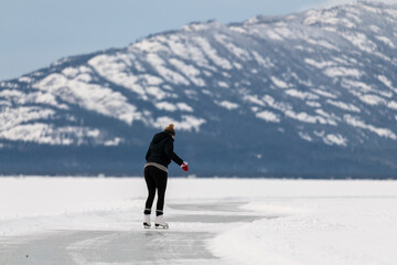 Fototapeta na wymiar Woman skating on frozen lake in northern Canada with huge mountains in the background surrounded by snow, snow capped and glassy ice on lakes surface. 