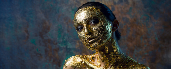 Girl with a mask on her face made of gold leaf. Gloomy studio portrait of a brunette on an abstract background.