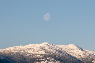 Full moon setting over the top of snow capped mountains in northern Canada during winter season. Woods, forest landscape below with perfect blue skies sky. 
