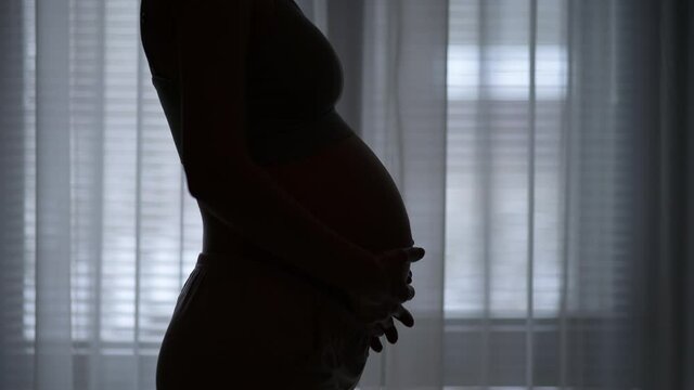Silhouette of a young pregnant slender woman appears on the background of the window in the evening. Woman stroking pregnant belly. Image of pregnancy and expectation of a child