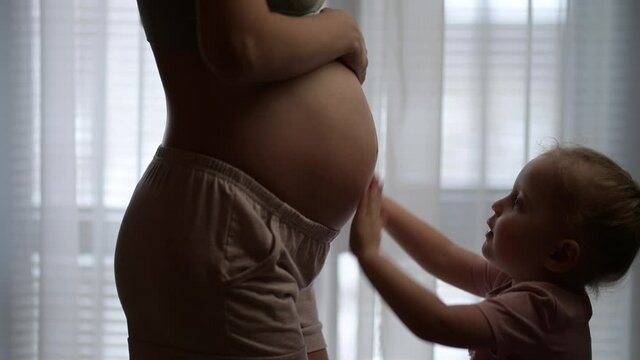 Little girl kissing her young mom's pregnant belly on a background of a window. Image of pregnancy and expectation of a child