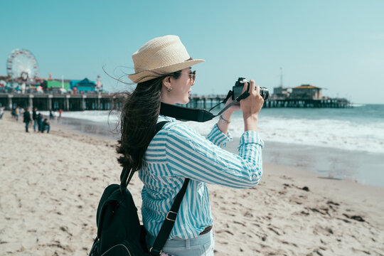 asian japanese woman backpacker having fun on beach with blue sea taking picture on camera. beautiful lady tourist wearing hat and sunglasses carrying backpack. female traveler enjoy ocean view.
