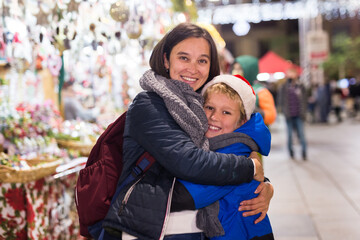 Fototapeta premium Portrait of happy woman hugging her cheerful preteen son during walk at traditional city Christmas fair