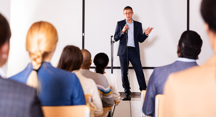 Confident lecturer standing with microphone on stage in conference room, speaking to businesspeople...