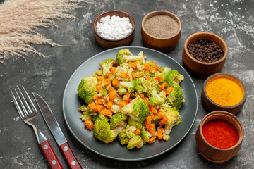 Top view of healthy meal with brocoli and carrots on a black plate and spices with fork and knife