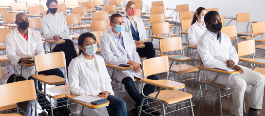 Group of people in white coats and protective masks sitting in conference room keeping distance at professional training for health workers. Precautions during mass events in coronavirus pandemic - obrazy, fototapety, plakaty