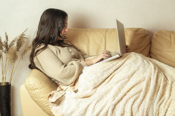 The girl sits on the sofa covered with a blanket and prints the text on the laptop.  Remote work online.  Student or schoolgirl on distance learning. Home work concept.