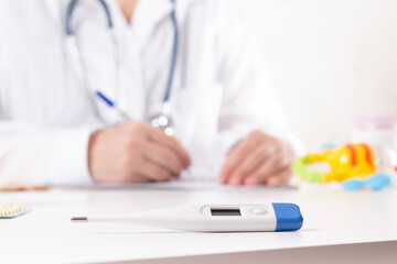 Pediatrician neonatologist writes pills prescription or diagnosis during visit in clinic . Electronic thermometer in front of blurred doctor in uniform and toys. Baby health care concept. 