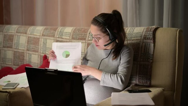 A young pregnant woman is holding a video communication conference, showing the printed graphs. Remote work from home via video communication