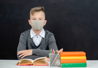 Teen boy wearing protective mask sits with books at school near blackboatd during corona virus and flu outbreak. Empty space for text