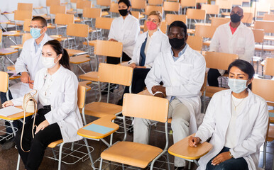 Fototapeta na wymiar Multiethnic group of young adult medicals in protective face masks attentively listening to lecture during training program for health workers, keeping social distance during pandemic situation