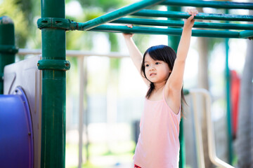 Young Asian girl hanging on monkey green bars in playground. Summer outdoor activity for child. Active preschool kid doing exercise sport. Happy healthy childhood.