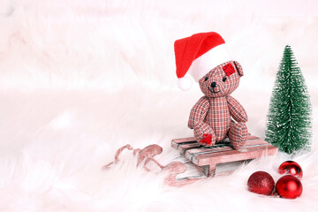 Little toy bear in a santa hat on a sleigh with fir tree on the white snow fur.