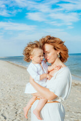 A young mother with red curly hair with her little daughter, a toddler in light clothes in the summer on the seashore, enjoy the rest. Happy family and children. Summer vacation and tourism
