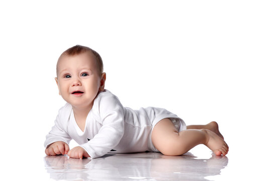 Upset infant baby boy toddler in diaper and white bodysuit with long sleeves is lying on floor on his stomach and going to cry, looks up with hope over white background. Infancy and babyhood concept