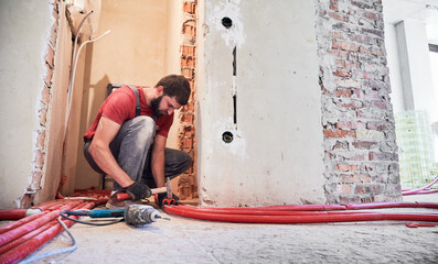 Man plumber in work overalls using hammer while laying pipes on the floor. Bearded male worker in...