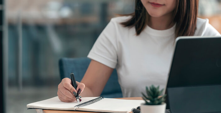 Closeup imange of female hand writing on notebook with pen while sitting at office desk.