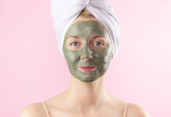 A young woman with a towel on her head and a clay mask on her face on a pink background. Face care, Spa treatments at home.