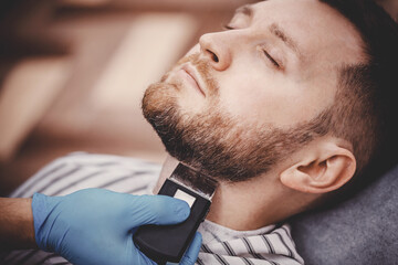 Obraz premium Close-up of barber with medical gloves shearing beard to man in barbershop. Haircut coronavirus pandemic protection