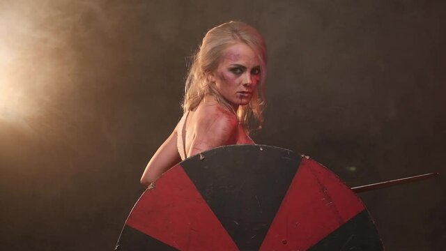 Nude and grimy barbaric woman from north with long gray hairs posing in dark background shaking a shield and changing a battle stances. Armed with sword and wielding a shield female warrior with
