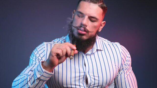 Strong and handsome bodybuilder with beard and moustache dressed in white shirt smokes a cigar posing in dark blue background. Stylish muscular guy with fashionable haircut smoking cigar.