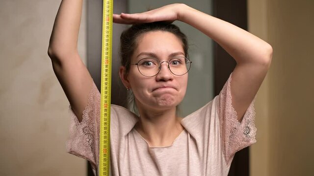 A young woman shows joy at her growth by holding a measuring tape next to her. The growth of a short woman and positive emotion