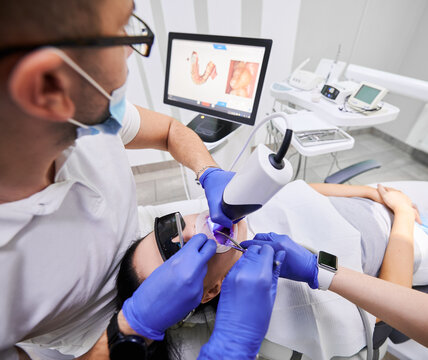 Dentist and his assistant scanning patient's teeth with modern scanning machine. Digital print of patient's teeth is on big screen. Modern high precision technologies. Concept of modern dentistry
