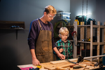 a young male carpenter is teaching woodwork to a young boy in his workshop