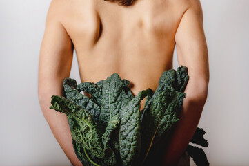 Vertical close-up unrecognizable naked back of beautiful woman. Lady holding a bunch of green leafy vegetable on white background. Vegan, diet, healthy lifestyle concept.