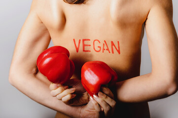 Close-up part of woman body naked back with red vegan write on the skin and holding two sweet peppers. Womanly, healthy lifestyle, diet, vegan and vegetarian concept.